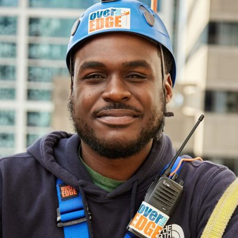 The image shows a smiling Black man wearing a blue helmet with the words "over EDGE" on it. He is also wearing a blue hoodie and has a walkie-talkie attached to his harness. The background is blurred, but it appears to be an urban setting with tall buildings.